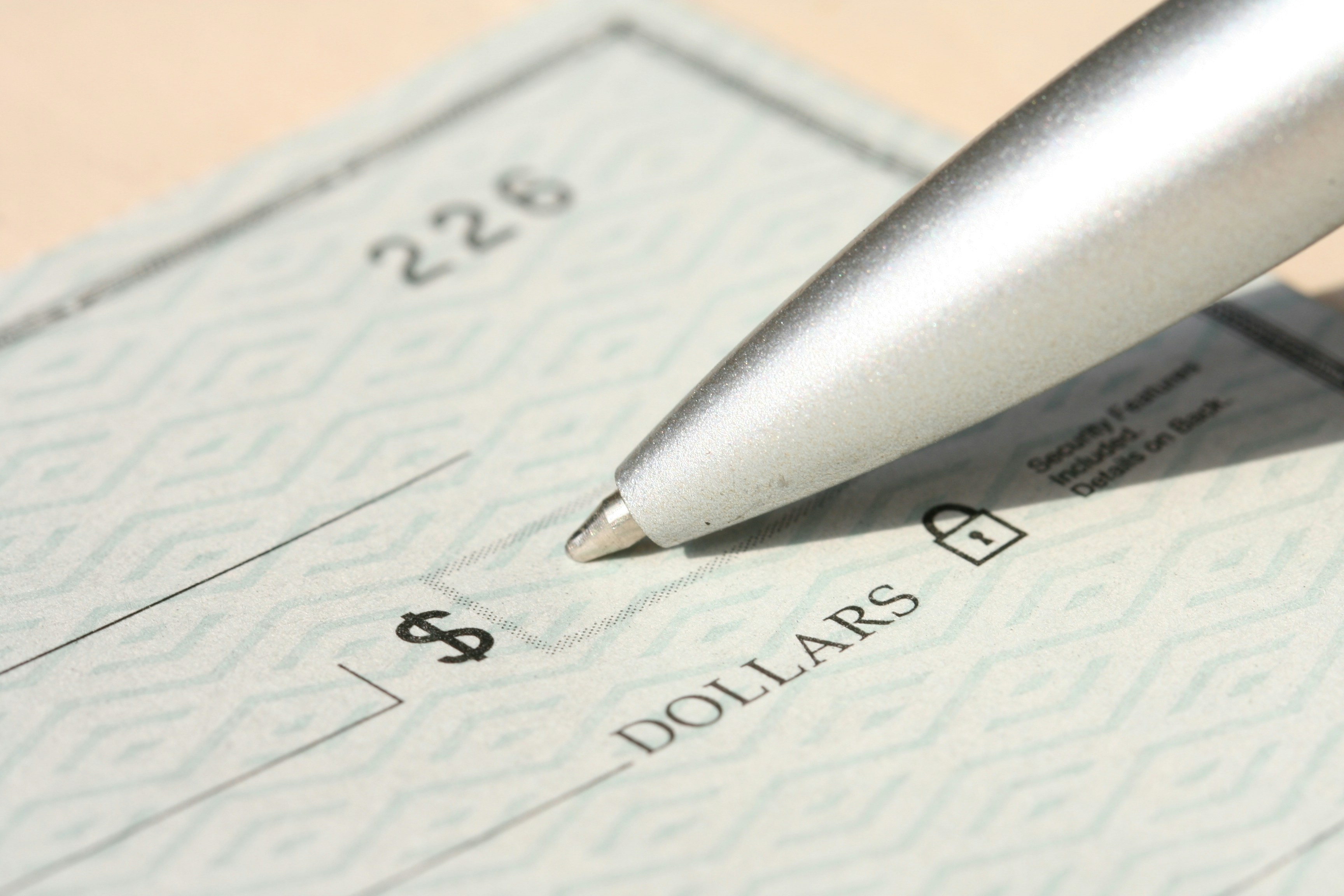 A close-up, high-resolution image of a blank check with a security pattern, showing the dollar sign box and part of the “DOLLARS” line. A metallic ballpoint pen rests near the writing area.